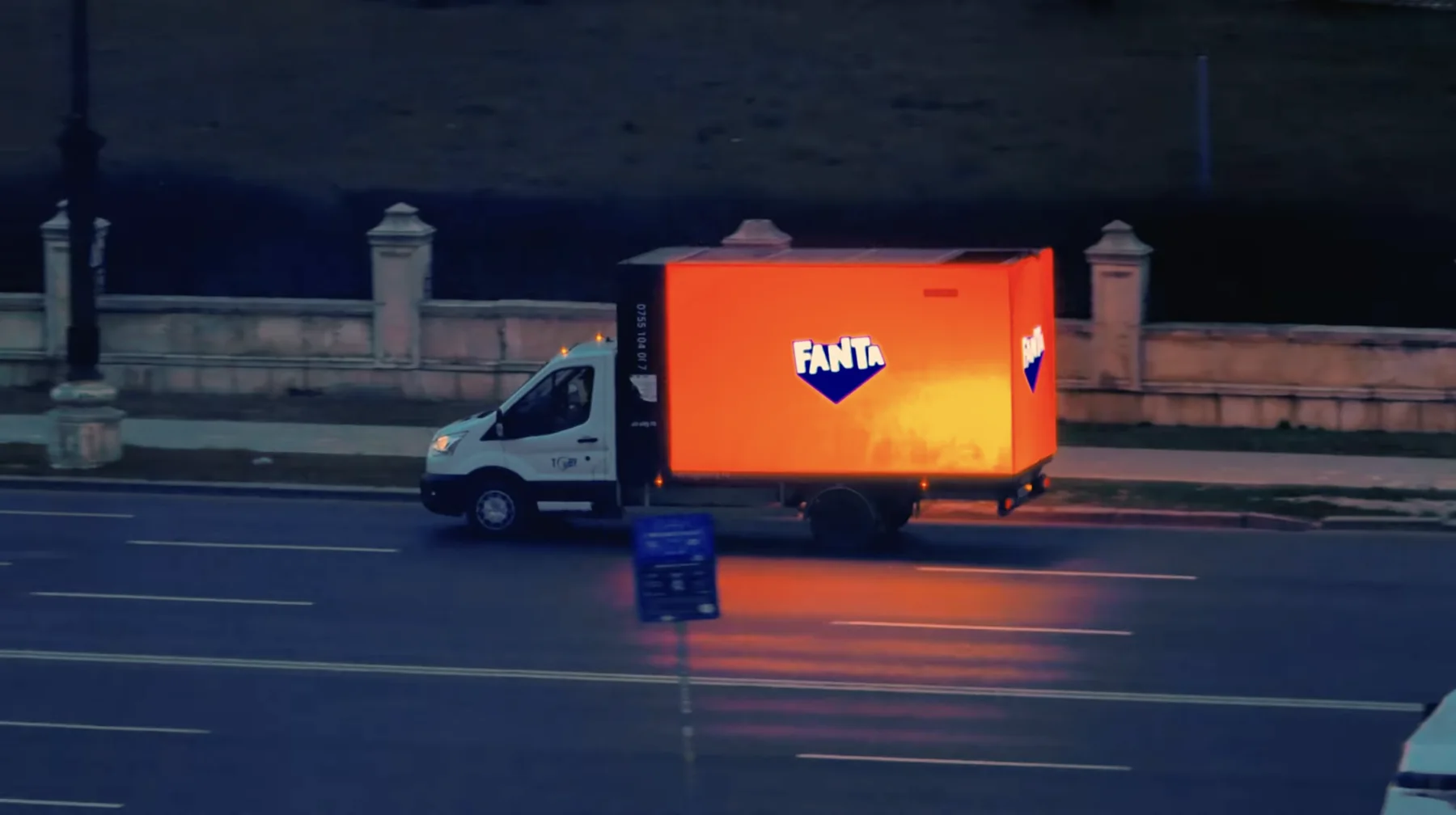 A Fanta truck with a glowing orange box driving past at dusk.
