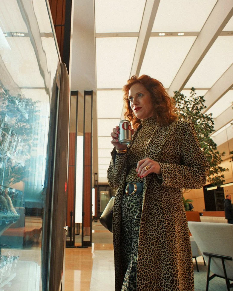 Film still: a woman in a leopard-print coat drinking Diet Coke beside a lit display case.